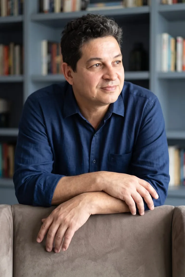 Dr. Carlos Fabiano de Oliveira, de camisa azul, apoiado no encosto de um sofá diante de uma estante de livros